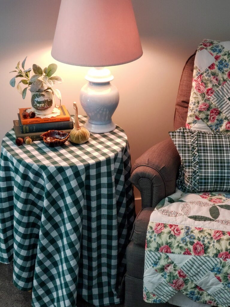 table with check tablecloth, lamp and stack of books. a quilt on a chair with a pillow.