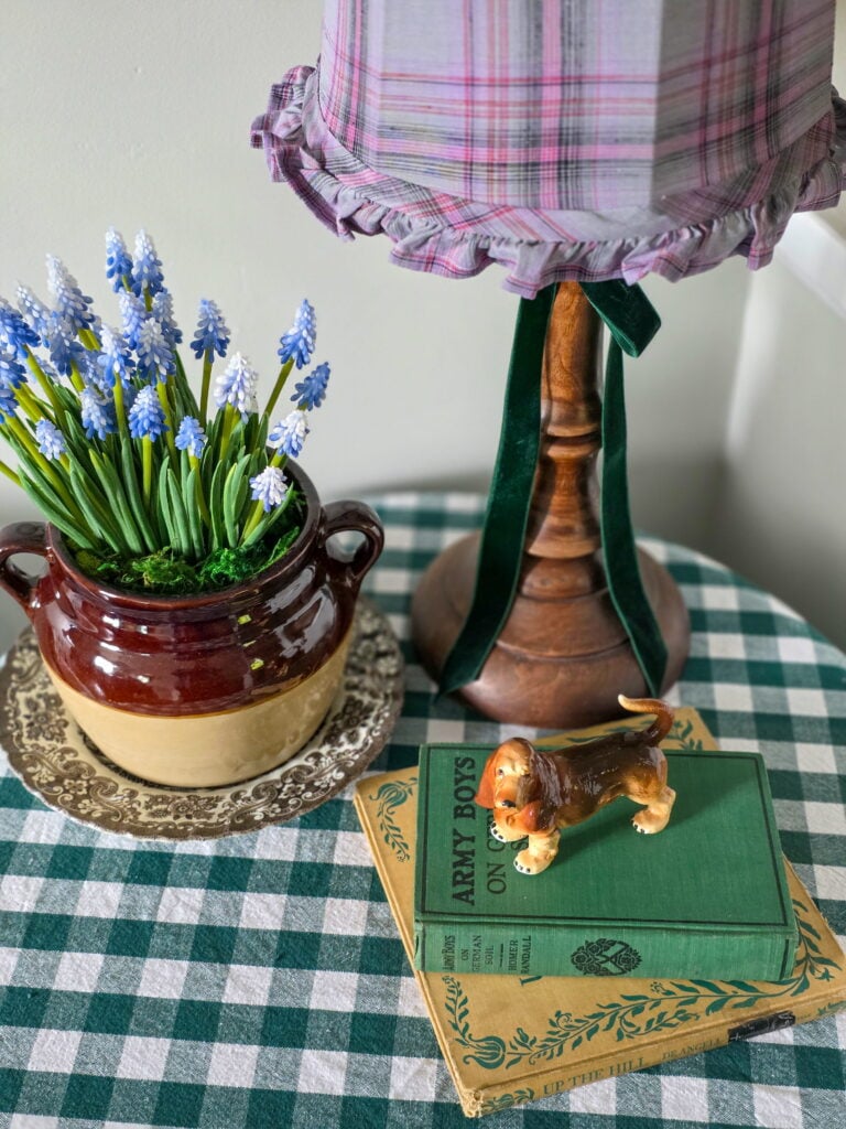 lamp with purple shade on gingham covered table with stack of books, basset hound object and planter with florals