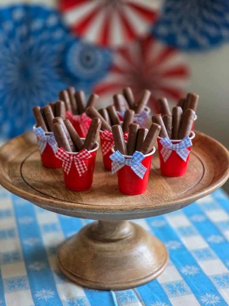 DIY vintage Americana party favor with chocolate raspberry sticks in mini red cups and gingham bow sitting on a wood cake stand