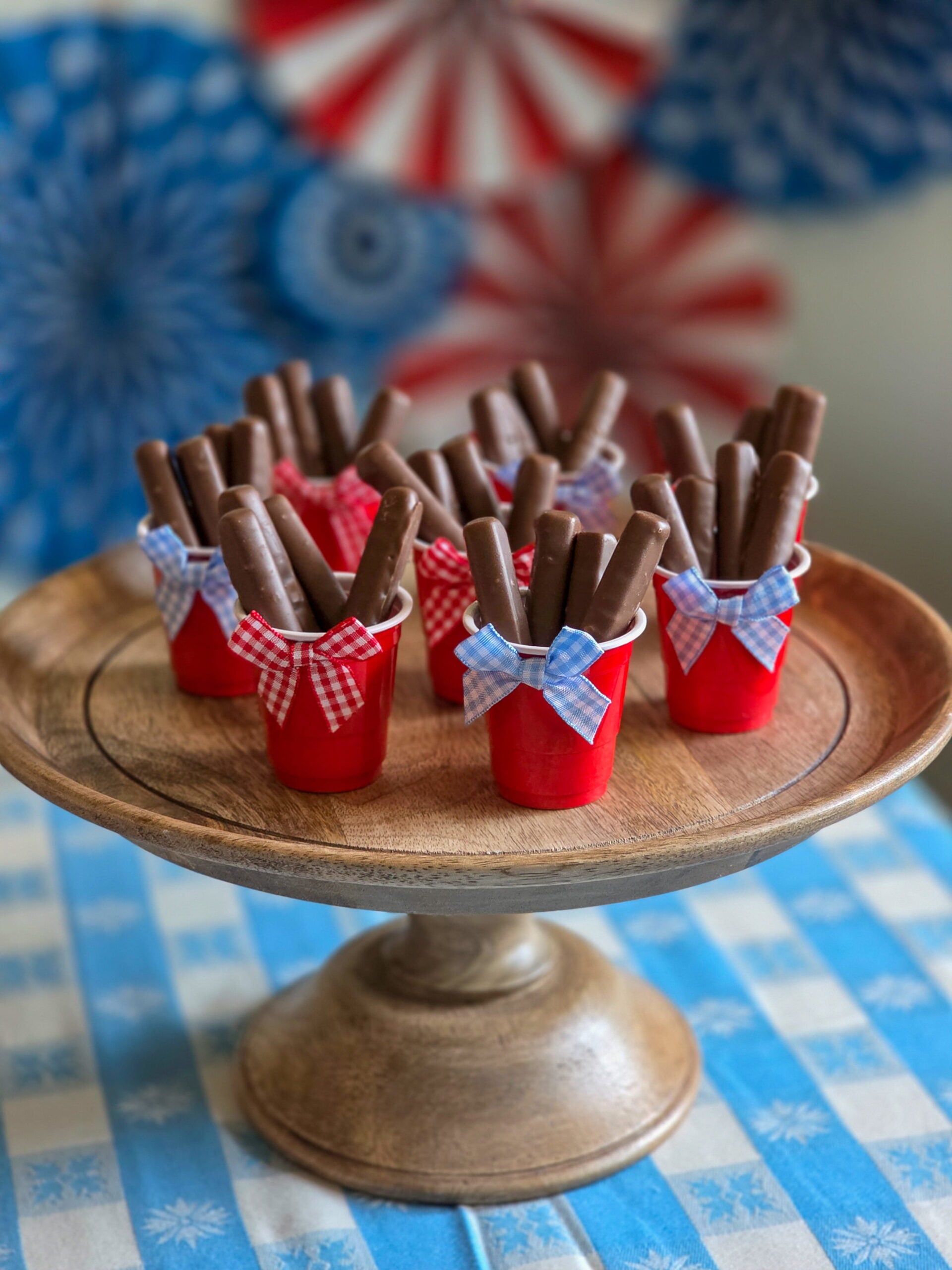 DIY vintage Americana party favor with chocolate raspberry sticks in mini red cups and gingham bow sitting on a wood cake stand