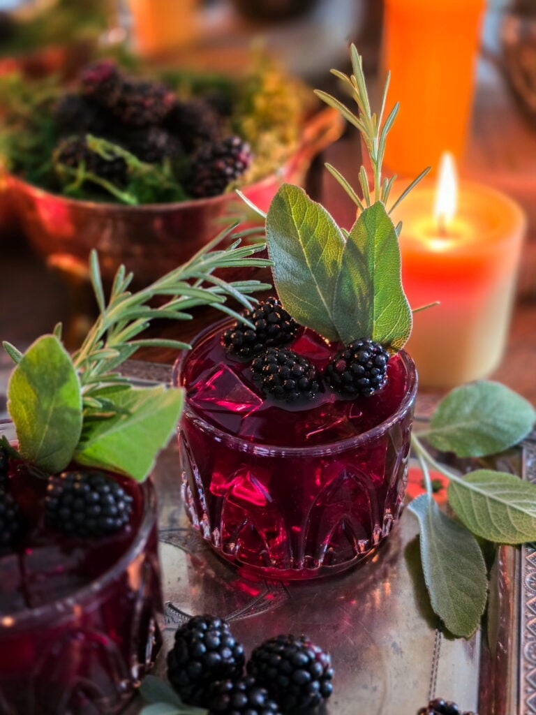 gillian owens blackberry bourbon cocktail with sage garnish sitting on vintage silver tray with blackberries and warm candles lit