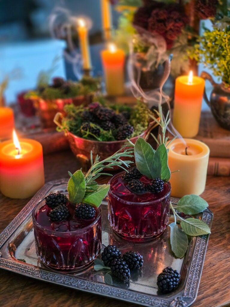 gillian owens blackberry bourbon cocktail with sage garnish sitting on vintage silver tray with blackberries and warm candles lit, and one candle blown out with smoke wafting