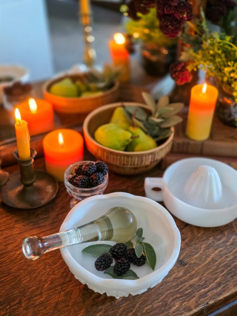 vintage glass pestle in milk glass bowl with blackberries and sage leaves for muddling. lit candles and a glass reamer, a bowl of bears and a bowl of blackberries