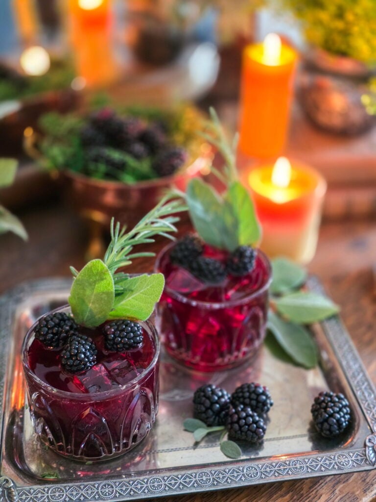 gillian owens blackberry bourbon cocktail with sage garnish sitting on vintage silver tray with blackberries and warm candles lit