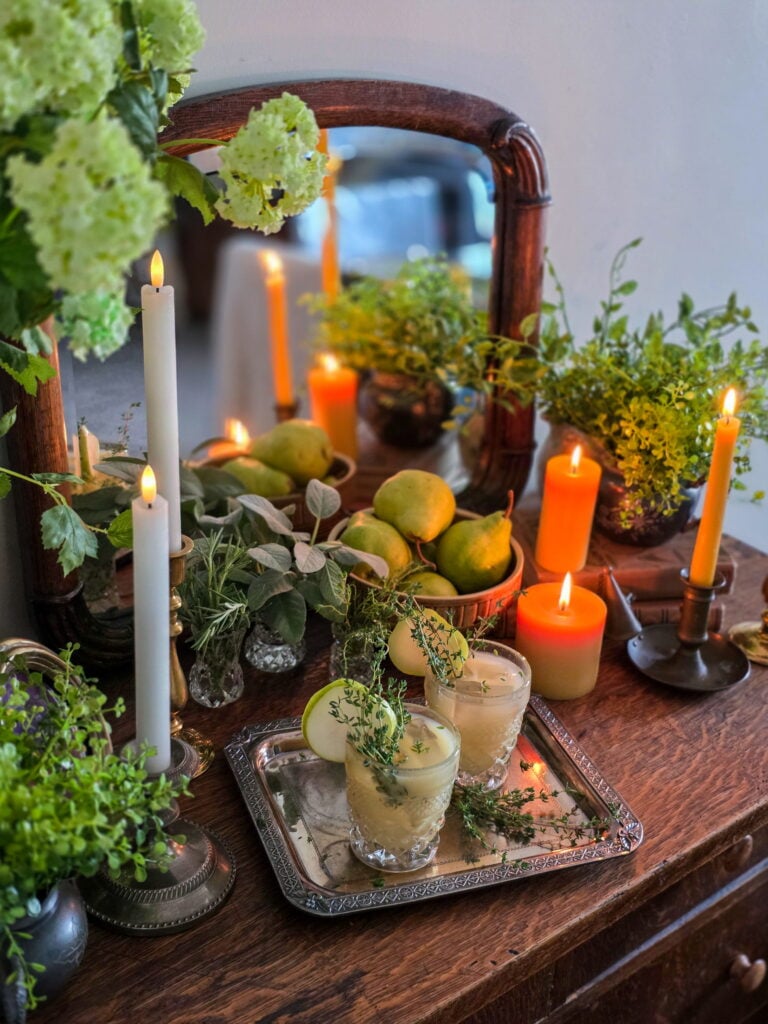 gin pear cocktail in vintage glass with thyme sprig garnish, and lit candles and a bowl of pears on a vintage wood bar