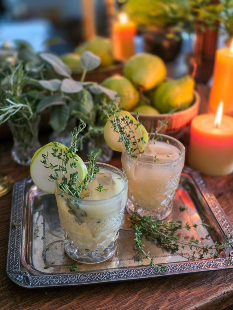 gin pear cocktail in vintage glass with thyme sprig garnish, and lit candles and a bowl of pears