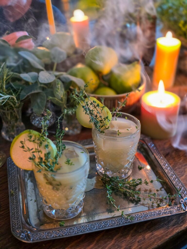 gin pear cocktail in vintage glass with thyme sprig garnish, and lit candles and a bowl of pears, and smoke wafting