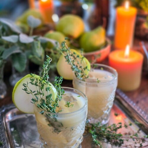 gin pear cocktail in vintage glass with thyme sprig garnish, and lit candles and a bowl of pears